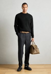 Pulover din tricot negru cu nervuri, curea bej, pantaloni elegant închiși la culoare, loafers negri, ținând o geantă din piele maro, stând pe podea din lemn.