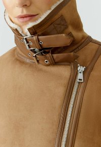 Veste en daim marron avec un col haut, doublure en fourrure, fermeture éclair argentée et sangles en cuir avec boucles en métal. Surface texturée.