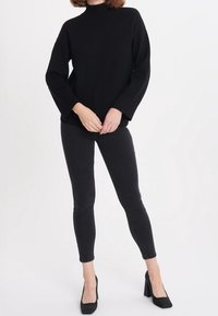 Czarny dziergany sweter z wysokim kołnierzem, oversize'owymi rękawami, zestawiony z czarnymi, dopasowanymi jeansami i czarnymi szpilkami.