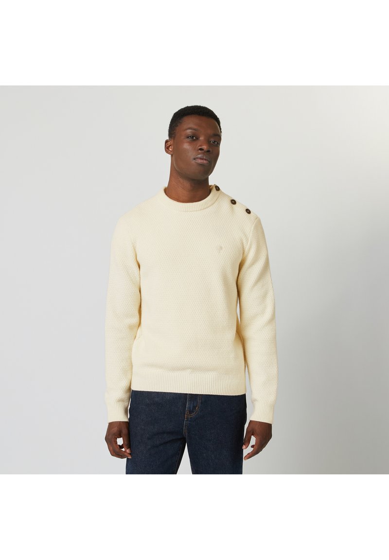 Pull en maille texturée crème avec un col rond et trois boutons marron sur l'épaule gauche. Porté avec un jean bleu marine.
