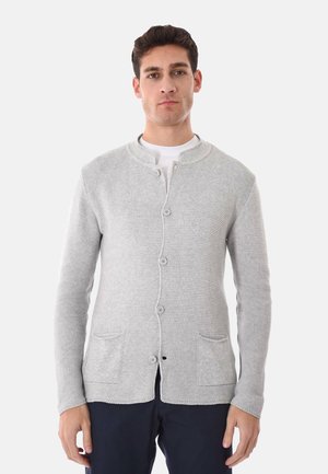 Uomo che indossa un cardigan grigio chiaro con bottoni, due tasche frontali e pantaloni scuri, rivolto verso lo spettatore su uno sfondo neutro.