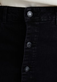Črna denim krilka z zapiranjem na gumb na sprednji strani, petimi vidnimi gumbi in gladkim, teksturiranim materialom z rahlimi podrobnostmi v šivanju.