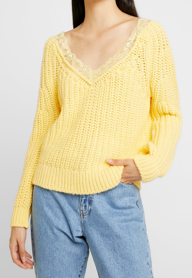 Femme portant un pull en maille jaune épais avec un col en V bordé de dentelle, associé à un jean bleu clair taille haute, une main dans la poche.