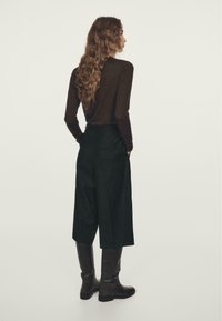 Donna con capelli lunghi e mossi che indossa una maglia a maniche lunghe marrone scuro, pantaloni neri a gamba larga e corti, e stivali neri al ginocchio, in piedi di profilo.