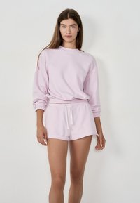 Młoda kobieta z długimi brązowymi włosami, ubrana w jasnofioletowy sweatshirt oraz dopasowane szorty na sznurkach, stojąca przed gładką białą ścianą.