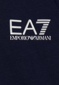 Dunkelblauer Stoff mit einem erhabenen, glänzenden weißen "EA7"-Logo und einem Akzent sowie "EMPORIO ARMANI" in kleinerer Schrift darunter.