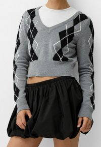 Grå kort tröja med svart och vit argyle-mönster, stickad textur och V-halsdesign, parat med en svart, rynkad kjol.