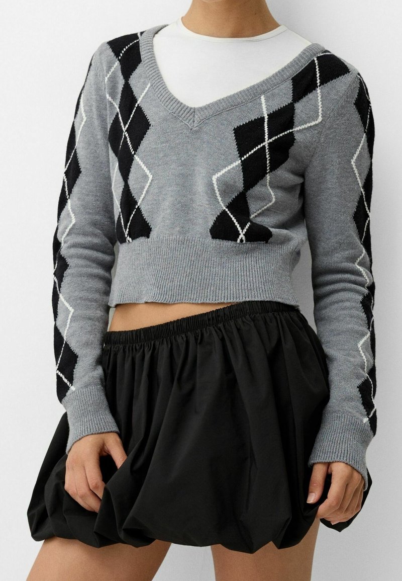 Grå kort tröja med svart och vit argyle-mönster, stickad textur och V-halsdesign, parat med en svart, rynkad kjol.