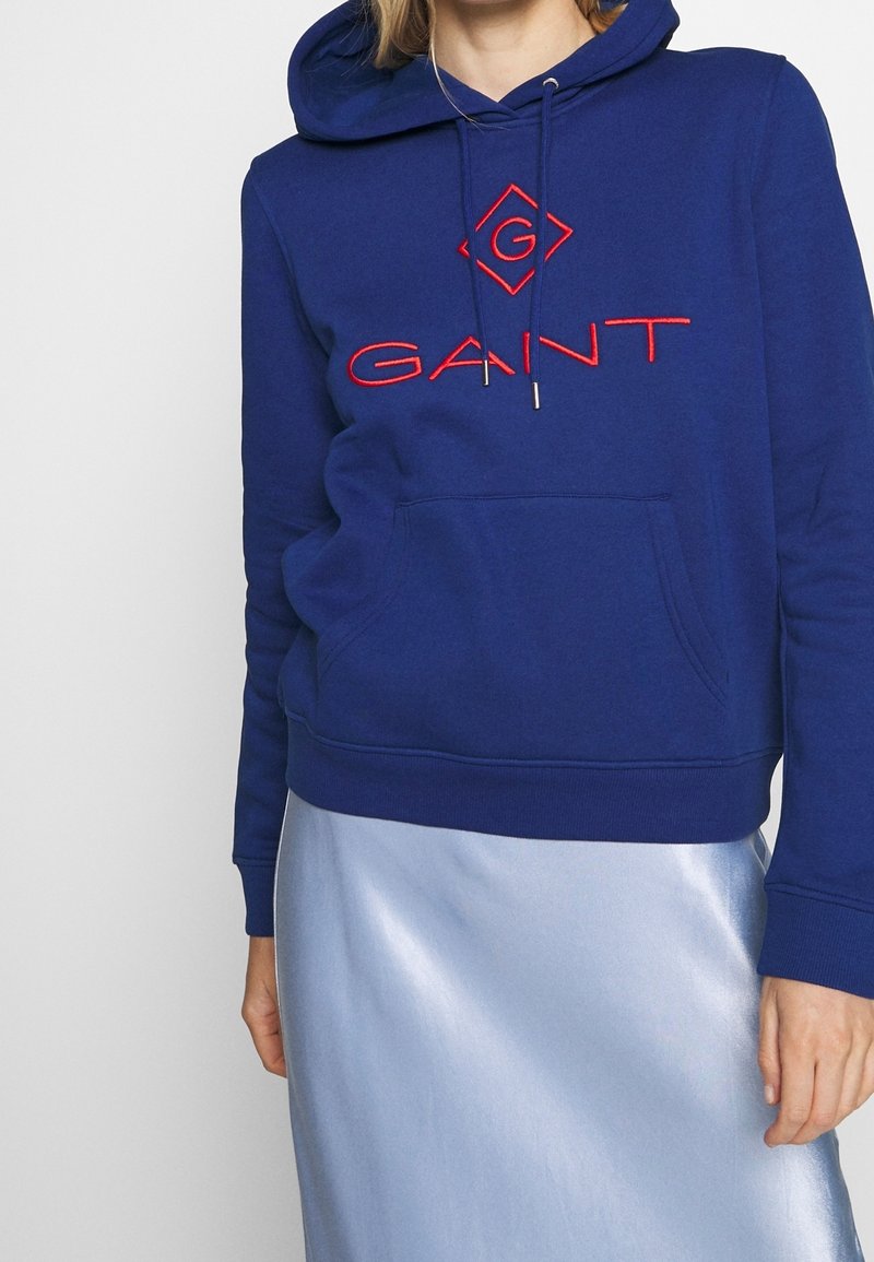 Personne portant un sweat à capuche GANT bleu marine avec un logo rouge et une jupe en satin bleu clair, debout devant un fond blanc uni.