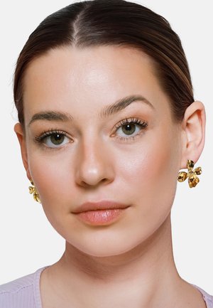 Kobieta o gładkiej skórze, brązowych włosach zaczesanych do tyłu, nosząca złote kolczyki w kształcie kwiatów oraz jasnofioletową bluzkę na gładkim tle.