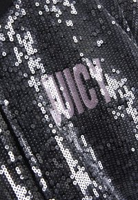 Fekete flitteres szövet, amely nagy, félig átlátszó rózsaszín "JUICY" feliratot tartalmaz. Az anyag fényes, sima felületű.