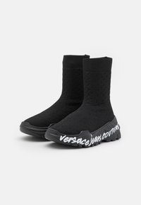 Botas de tobillo de punto negras con parte superior texturizada y suela de goma gruesa que presenta la letra blanca "versace jeans couture" a lo largo del costado.