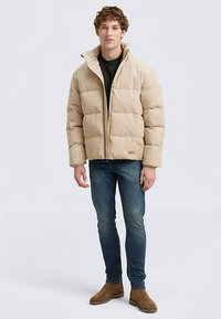 Beige puffjacka med dragkedja framtill, med fyrkantiga, vadderade sektioner. Bärs över en svart skjorta, matchas med blå jeans och bruna stövlar.