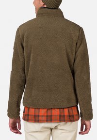 Giacca in pile verde oliva con superficie texturizzata, colletto alto e taglio dritto, indossata sopra una camicia a quadri rossi e neri.