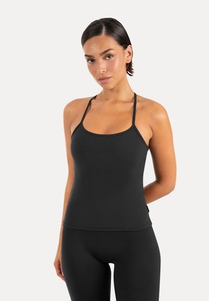 Kvinde med kort mørkt hår iført en sort tætsiddende tanktop med tynde stropper og sorte leggings mod en ensartet lys baggrund.