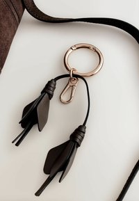 Kulcstartó kör alakú fém gyűrűvel, fekete bőrszálakkal és kis fém kapoccsal; elegáns anyag- és textúra-kontrasztokkal rendelkezik.