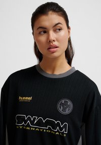 Zwart sportshirt met lange mouwen en subtiele streepjes, grijze kraag en gele logo-accenten met de tekst "hummel" en "SWARM." Bevat een ronde embleem.