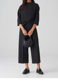 Femme portant un haut texturé noir et un pantalon large, tenant un sac à main matelassé noir et portant des mocassins noirs à talon épais.
