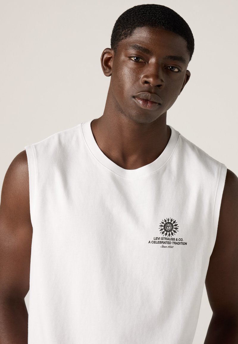 Jonge man met kort haar die een wit mouwloos Levi Strauss-shirt draagt met een klein zwart zonlogo en tekst op de borst.