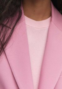 Gros plan d'une personne portant un blazer rose clair sur un pull rose pâle, avec de longs cheveux foncés visibles sur le côté gauche.