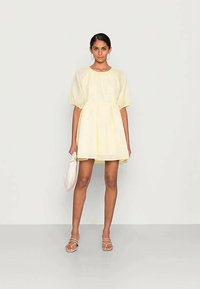 Mulher vestindo um vestido curto amarelo claro com mangas bufantes, segurando uma bolsa de mão branca e usando sandálias brancas com tiras, em pé diante de uma parede simples.