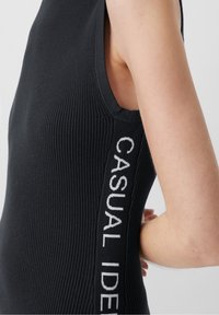 Vêtement sans manches noir à côtes avec un grand texte "CASUAL IDEAS" sur le côté en blanc. Design ajusté avec une surface texturée.