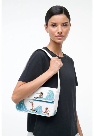 Femme en chemise noire tenant un sac d'épaule blanc avec des surfeurs brodés colorés chevauchant des vagues bleues.