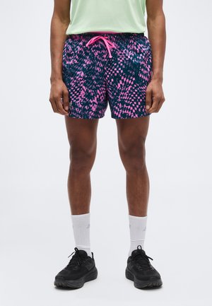 Férfi, tengerészkék és rózsaszín mintás rövidnadrágot visel, rózsaszín húzózsinórral, fehér bokazokniban, fekete sportcipőben és világoszöld ingben áll.