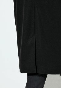 Cappotto lungo nero con una texture liscia, dotato di tasche laterali e uno spacco laterale. Abbinato a jeans scuri, visibile dalla vita in giù.