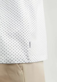 Fehér ing kis sötétkék geometriai mintával, oldalán címkével. Az anyag simának és könnyűnek tűnik.