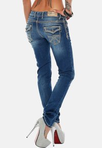 Jean en denim bleu ajusté avec des coutures décoratives, deux poches arrière et une étiquette en cuir. Assortis avec des escarpins argentés.