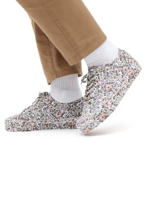 Personne portant des chaussettes blanches et un pantalon beige avec des baskets blanches à motifs floraux roses et verts.