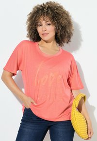 Mujer con cabello rizado, vistiendo una camiseta coral rosa con la frase "Love Out Loud" y jeans oscuros, sosteniendo un bolso amarillo de mimbre frente a un fondo blanco.