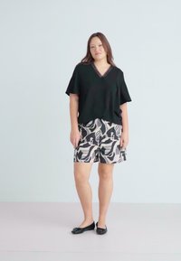 Musta V-kaula-aukkoinen toppi lyhyillä hihoilla, yhdessä kuviollisten beige-shortsien kanssa, joissa on abstrakteja kuvioita. Yhdistetty mustiin litteisiin kenkiin.