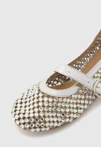 Scarpa piatta beige con rete trasparente ricoperta di rotondi strass argentati e una fascia in pelle verniciata bianca con chiusura a fibbia.