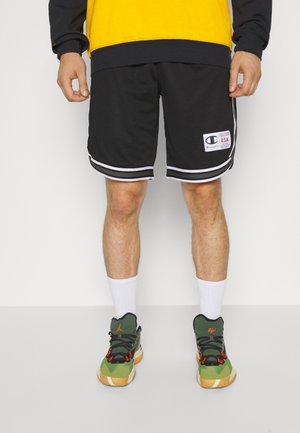 Černé basketbalové kraťasy z průsvitné síťoviny s bílými pruhy po stranách a nášivkou loga Champion na pravé noze; nošeny se zelenými vzorovanými teniskami a bílými ponožkami.