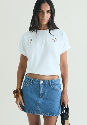 Kobieta w białym skróconym T-shircie z wycięciami i niebieską dżinsową spódnicą, trzymająca małą plecioną torebkę i nosząca masywne bransoletki.