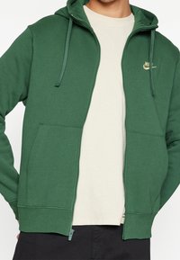 Grön zip-up hoodie i mjukt tyg, med en framficka, justerbar dragsko på huven och en guld Nike-logotyp på vänster bröst.