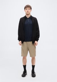 Μαύρο τζάκετ με φερμουάρ πάνω από μπλε μπλουζάκι πόλο, καφέ cargo σορτς, μαύρες κάλτσες και μαύρα παπούτσια. Ομαλή υφή υφάσματος, πρακτικός σχεδιασμός με τσέπες.