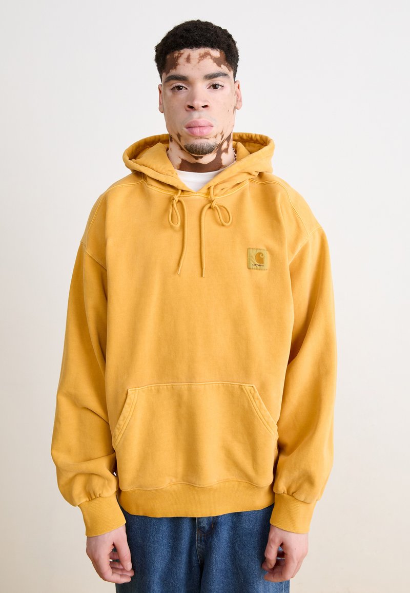 Hoodie en coton jaune avec une poche frontale, un capuche à cordon et des poignets côtelés. Présente un petit écusson logo sur la poitrine. Porté avec un jean bleu.