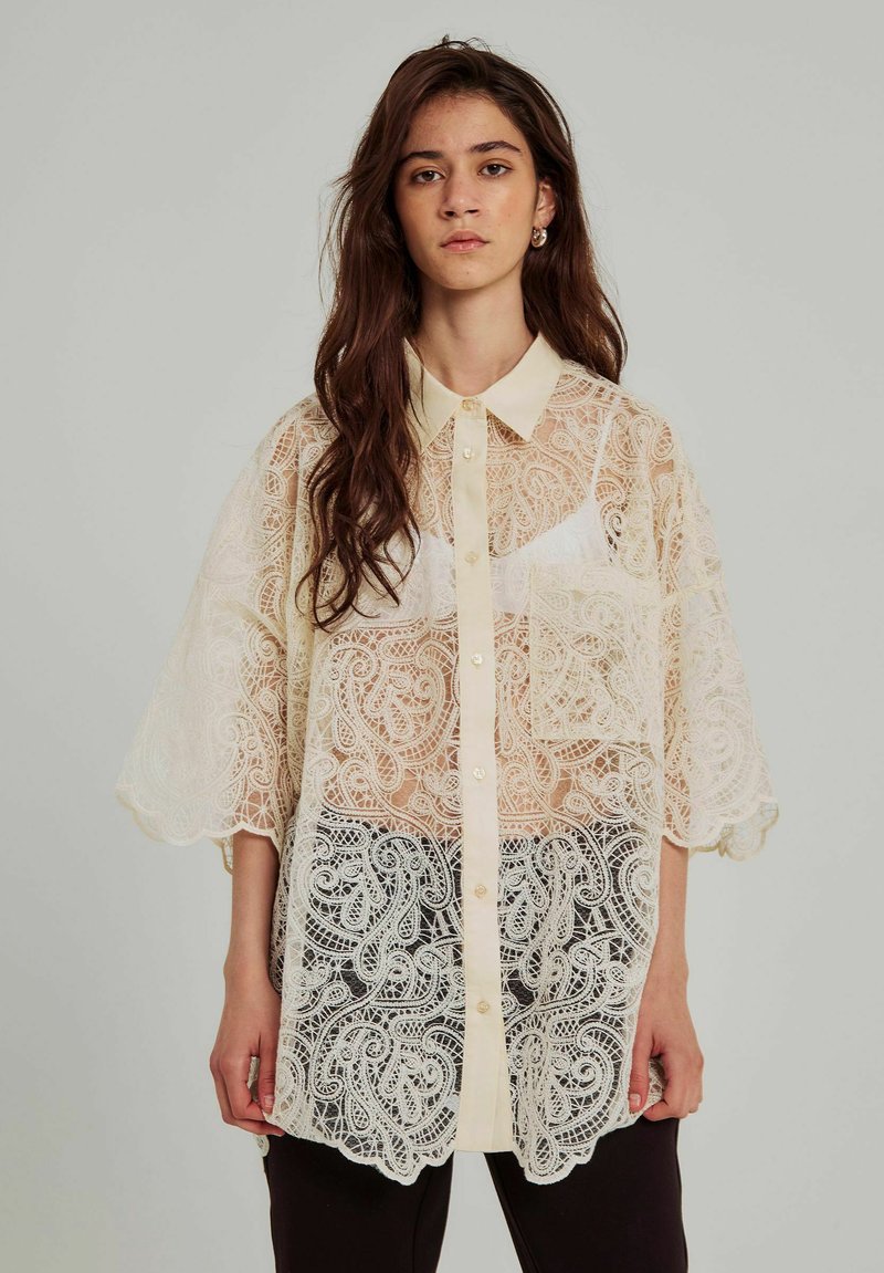 Blusa in crema con sovrapposizione di pizzo, colletto, maniche corte, chiusura a bottoni e intricati motivi paisley sull'orlo.
