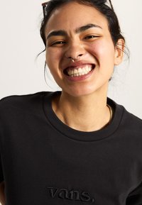 Camiseta de algodón negra con cuello redondo que presenta el logo bordado "vans." en el pecho, textura suave y corte relajado.