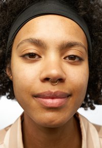 Zbliżenie na osobę z ciemnym opaską na głowie, kręconymi włosami, złotą kolczykiem w septum i naturalną teksturą skóry, ubrana w jasnobeżową koszulę w paski.