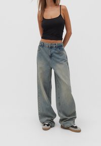 Frau, die ein schwarzes Träger-Crop-Top, weit geschnittene, hellblaue Jeans und schwarz-weiße Turnschuhe trägt und vor einem einfarbigen Hintergrund steht.