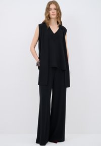 Débardeur noir sans manches avec un col en V et des détails texturés, associé à un pantalon noir à jambe large. Tissu lisse avec une silhouette fluide.