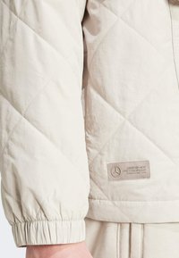 Svijetlosmeđa quilted jakna s teksturiranim završetkom. Sadrži elastične rukave i logotip s urezanim tekstom na lijevoj strani.