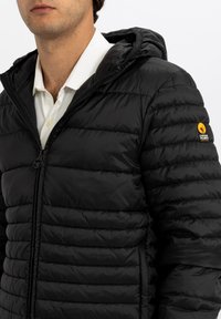 Giacca a piumino nera con chiusura frontale con zip, design trapuntato e cappuccio. Presenta un logo sulla manica sinistra, con una texture liscia e una finitura lucida.