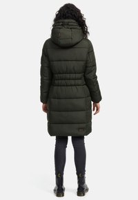 Dlhý, polstrovaný zelený kabát s kapucňou, stiahnutým pásom, vodorovným prešívaným vzorom a logom na spodnom leme. Nosený s tmavými nohavicami a čižmami.