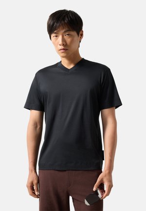 Jonge man die een zwart V-hals T-shirt en een bruine broek draagt, met een zwarte zonnebril tegen een lichtgrijze achtergrond.