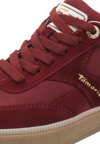 Κοντινό πλάνο κόκκινου σουέδ sneaker με χοντρά κορδόνια, ραμμένες λεπτομέρειες, μπεζ σόλα και χρυσή κεντημένη επωνυμία στη πλευρά.